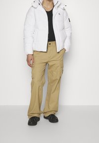 Giacca a vento bianca con zip e cappuccio, t-shirt nera, pantaloni cargo beige con tasche e scarpe da ginnastica nere. Design semplice, vestibilità casual.