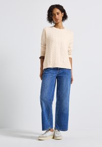 Nainen kermaisen värisessä palmikkoneuletakissa, sinisissä leveälahkeisissa farkuissa ja valkoisissa tennareissa, seisoo rentona valkoista taustaa vasten.