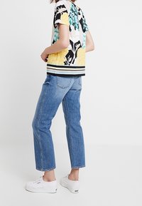 Korte mouwen bloemen top met gele, teal en zwarte patronen; gecombineerd met lichtblauwe rechte jeans en witte sneakers.