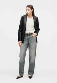 Γυναίκα όρθια, φοράει μαύρο δερμάτινο σακάκι, λευκό πουκάμισο με μακριά μανίκια, φαρδιά γκρι τζιν με μαύρη ζώνη και μαύρα μυτερά παπούτσια, κοιτάζει πλάγια.