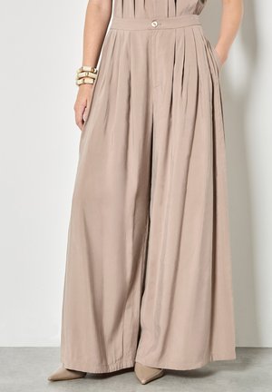 Kobieta w beżowych plisowanych spodniach o szerokich nogawkach, włożonym do środka topie, beżowych, szpiczastych butach i masywnych kremowych bransoletkach na lewej ręce.