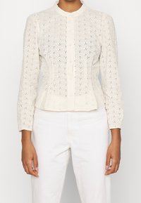 Hellbeige gestickte Bluse mit rundem Kragen, langen Ärmeln, Knopfverschluss und aufwendigen floralen Cut-out-Mustern. Glatte Textur.