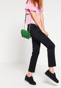 Top corto rosa a maniche corte con dettagli arricciati, pantaloni neri in tessuto testurizzato e una piccola borsa a tracolla verde. Sneakers nere chunky completano il look.