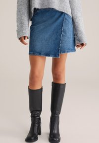 Denim obleka z zavihkom v srednje modri barvi, z razfrcanim robom in stranskim gumbom. V kombinaciji z črnimi usnjenimi škornji do kolen.