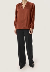Blusa ruggine a maniche lunghe con scollo a V, cuciture delle spalle arricciate e una superficie liscia, abbinata a pantaloni neri a gamba larga e scarpe a punta.