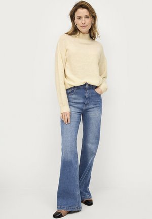 Maglione beige a maglia con colletto alto abbinato a jeans blu a campana. I jeans hanno una finitura sbiadita e l'outfit è completato con scarpe nere.