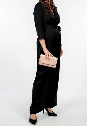 Femme en robe en satin noire et talons, tenant une pochette rose clair, debout devant un fond blanc.