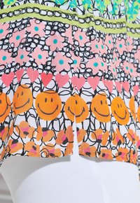 Motif de tissu coloré avec des fleurs roses, des visages souriants orange, des cœurs rouges et des lignes noires abstraites sur fond blanc au-dessus d’un pantalon blanc.