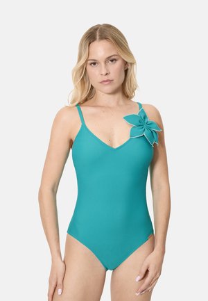 Mujer con un traje de baño entero color verde azulado con tirantes finos y una gran flor de tela adjunta cerca del hombro derecho.