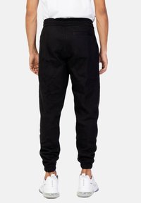 Persona vestida con pantalones jogger negros y zapatillas blancas de pie de espaldas a la cámara frente a un fondo liso.