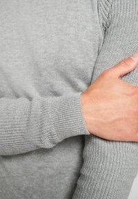 Světle šedý pletený svetr s texturovaným vzorem, se zpevněnými manžetami a měkkou tkaninou. Záběr na ruku opřenou o rukáv svetro.