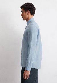 Lichtblauw, lang mouw, overhemd met knopen gemaakt van een textuurstof. Heeft een klassieke kraag en een geplooide achterkant, gecombineerd met donkere broek.