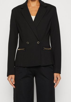Blazer noir sur mesure avec détail de chaîne dorée sur les poches, porté avec un pantalon noir assorti.