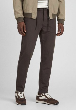 Mand iført mørkebrune slim fit bukser, beige jakke, stribet skjorte og brune og hvide sneakers, stående mod en ensfarvet baggrund.
