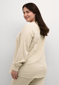 Beige Langarmshirt aus strukturiertem Stoff, mit lockerer Passform und gerafften Bündchen, kombiniert mit passenden beigen Hosen.