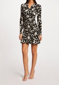 Czarna i biała sukienka w wzory, z długimi rękawami, dekoltem w kształcie litery V, dopasowaną talią i rozkloszowanym dołem. Noszona z cielistymi sandałami na obcasie.