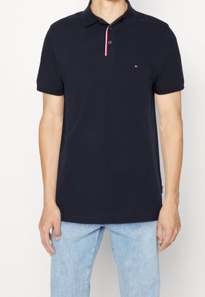 Homme portant un polo bleu marine à manches courtes avec un petit logo rouge, blanc et bleu sur la poitrine et un jean bleu clair.