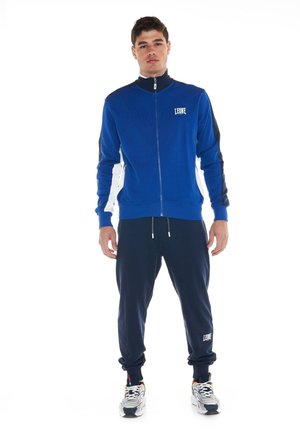 Un mannequin masculin se tient devant un fond blanc, vêtu d’une veste de survêtement bleue zippée, d’un pantalon de jogging bleu marine et de baskets blanches.