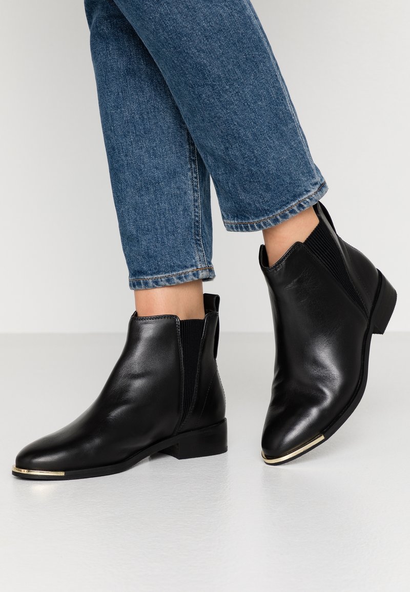 Bottes cheville en cuir noir avec des panneaux latéraux élastiques, bouts pointus et un accent doré sur le bout. Portées avec un jean bleu raccourci.