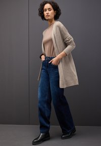 Dlouhý béžový kardigan z měkkého úpletu, nošený přes přiléhavý hnědý top. V kombinaci s tmavomodrými kalhotami s širokými nohavicemi a černými kotníkovými botami.