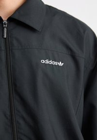 Black zip-up jacket made of smooth fabric. Features a collar and an embroidered white Adidas logo with a trefoil design on the left chest.