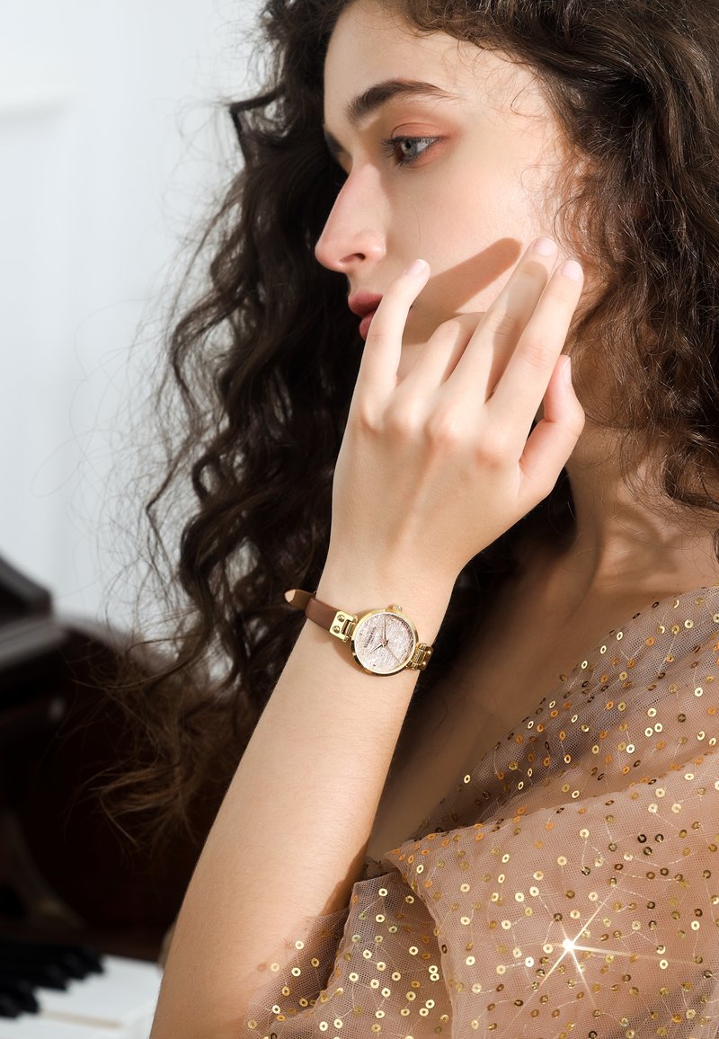 Montre dorée avec un cadran rond et texturé, bracelet en cuir brun et accents décoratifs. Modèle portant un haut transparent et pailleté.
