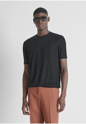 Hombre con gafas de sol negras grandes, camiseta negra de manga corta acanalada y pantalones de color óxido, posando contra un fondo claro y liso.