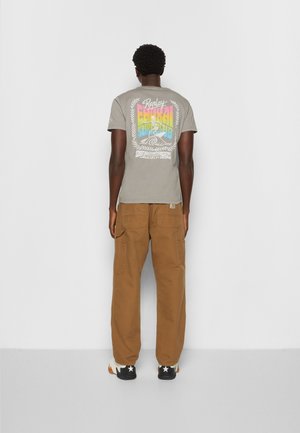 Homme portant un t-shirt graphique gris avec un design coloré de surf, un pantalon cargo marron et des baskets noires avec des étoiles blanches, debout face au mur.