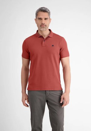 Man standing facing forward wearing a rust-colored polo shirt and gray pants against a plain white background.