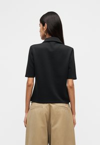 Μαύρο πόλο με κοντό μανίκι και γιακά, κατασκευασμένο από λεία υλικά. Διαθέτει χαλαρή εφαρμογή και ελαφρώς κυρτή κάτω ραφή.