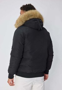 Veste noire matelassée avec capuche doublée en fausse fourrure, poignets et ourlet côtelés sombres, et poches latérales, vue de dos.