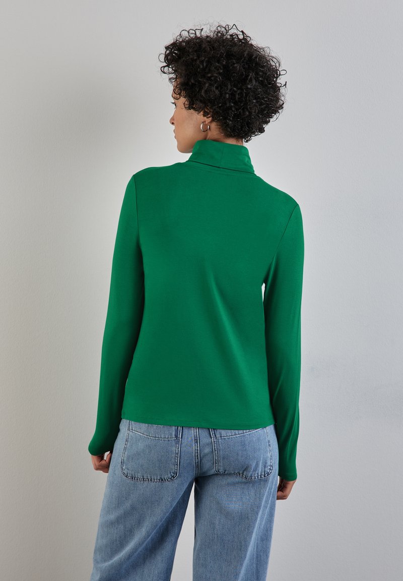 Zelený dlouhý rukáv rolák vyrobený z hladké tkaniny, na ramenou těsný a mírně volnější v pase, doplněný o světle modré džíny.