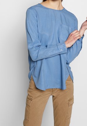 Personne portant une chemise ample bleu clair à manches longues avec une poche poitrine et un pantalon cargo beige, ajustant le poignet de la manche gauche.