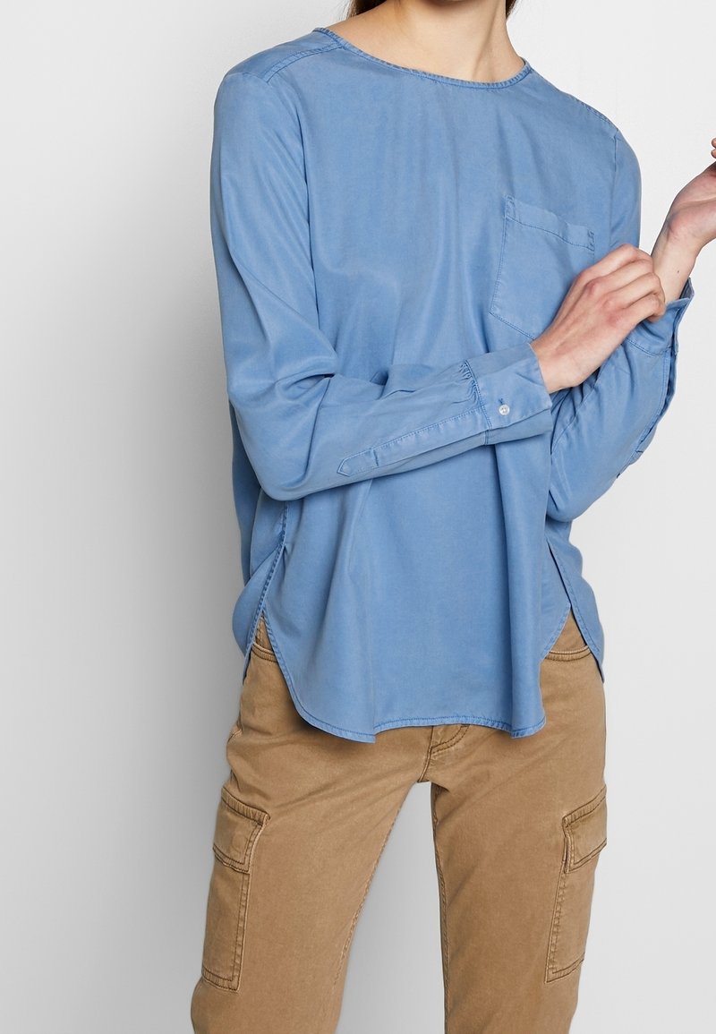Personne portant une chemise ample bleu clair à manches longues avec une poche poitrine et un pantalon cargo beige, ajustant le poignet de la manche gauche.