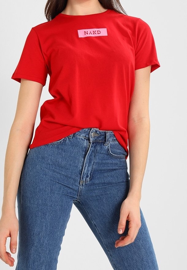 Femme portant un t-shirt rouge à manches courtes avec le logo "NAKD" et un jean bleu taille haute sur un fond clair uni.