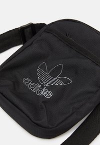 Bolso de hombro de tela negra con una correa ajustable, que presenta la marca blanca de Adidas y un logo gráfico. Textura suave, diseño compacto.