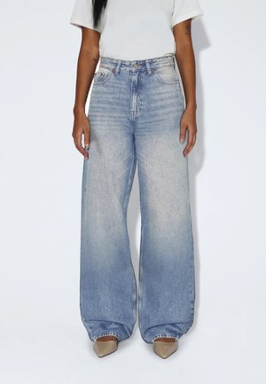 Person wearing high-waisted, loose light blue denim jeans and beige pointed shoes against a plain white background.