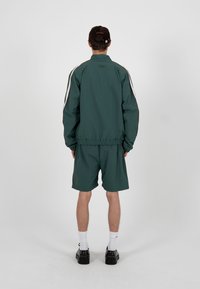 Homme portant une veste de survêtement vert foncé et un short, des chaussettes blanches, des chaussures noires et une casquette noire, debout de dos sur un fond uni.