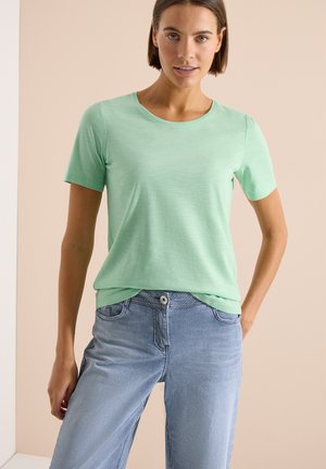 Femme portant une chemise manches courtes vert clair et un pantalon rayé bleu et blanc, debout devant un fond beige.