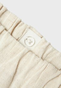 Tejido beige con un tejido texturizado, con una cinturilla fruncida y un detalle de botón transparente unido a un parche de tela blanca.