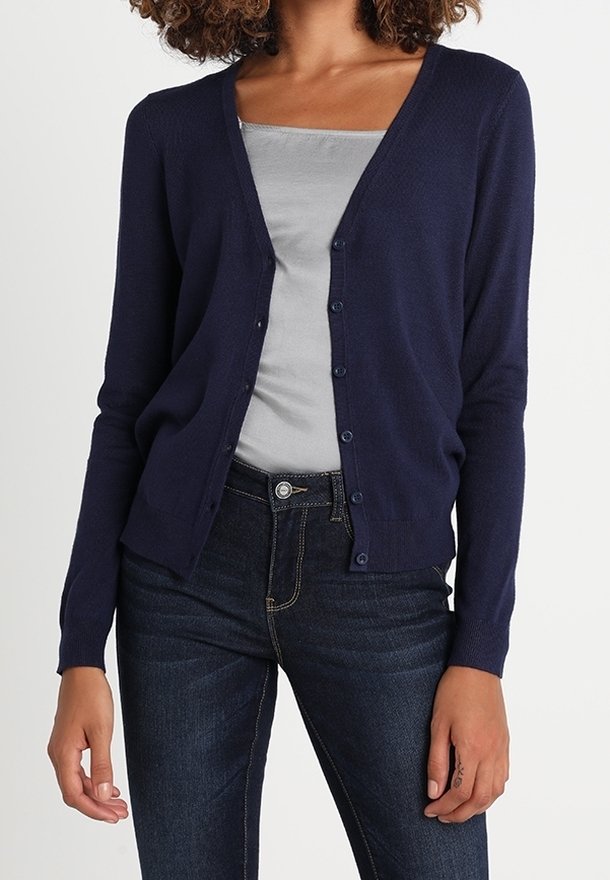 Femme portant un cardigan bleu marine boutonné par-dessus un débardeur gris clair et un jean bleu foncé, debout devant un fond blanc.