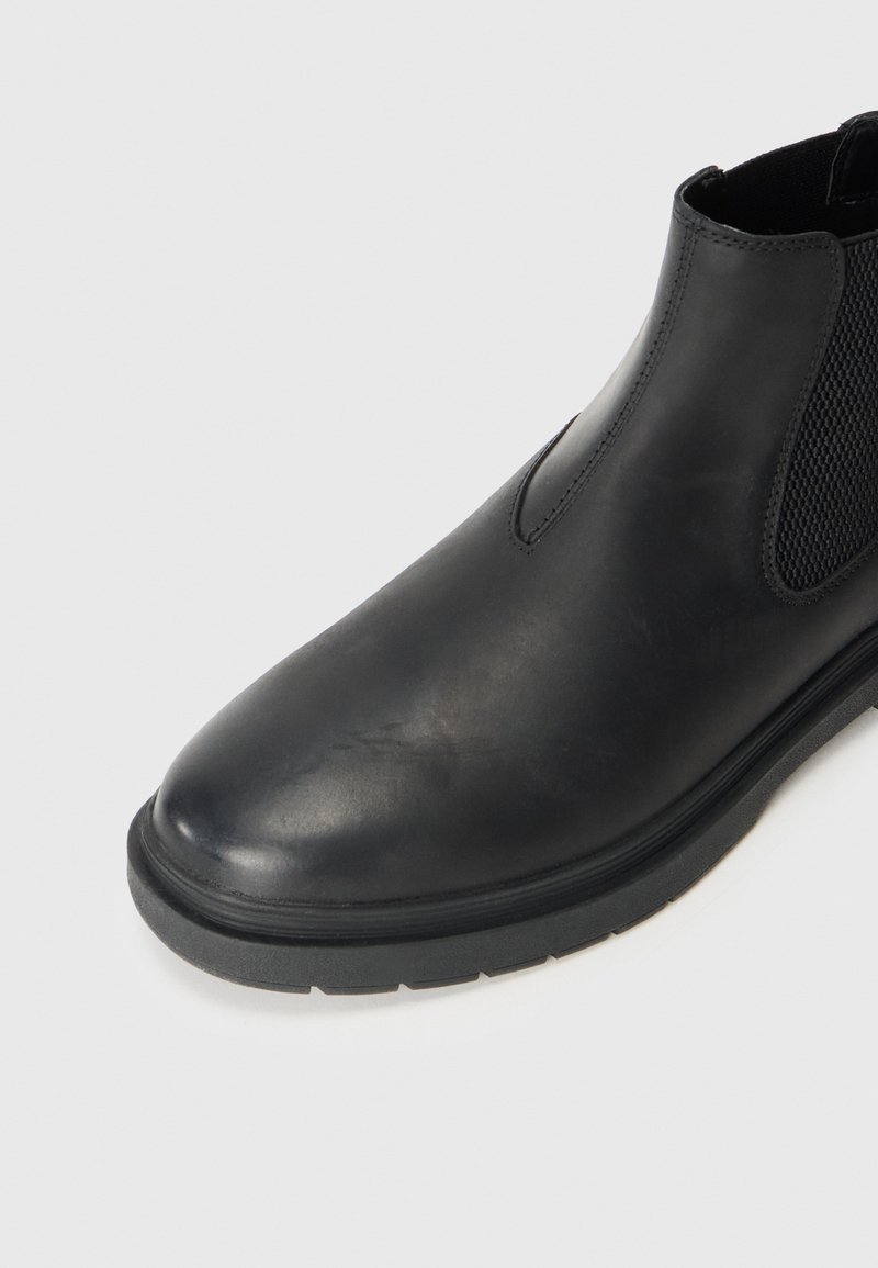 Bottines Chelsea noires en cuir lisse, avec des panneaux latéraux élastiques et une semelle en caoutchouc texturée. Bout rond avec un léger éclat.