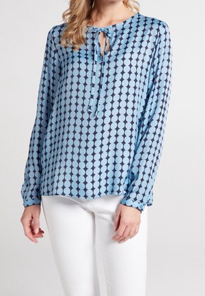 Vrouw die een lange mouwen blauwe blouse draagt met een groot polkadotpatroon en een strik bij de hals, gecombineerd met witte broek.