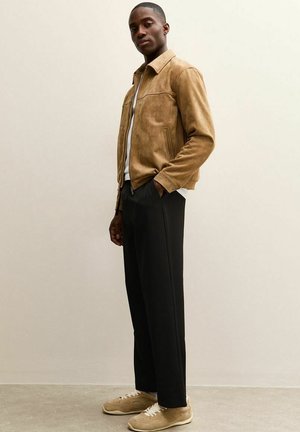 Giacca in camoscio color nocciola con zip frontale e tasca sul petto, camicia bianca sotto, abbinata a pantaloni neri e sneakers color nocciola.