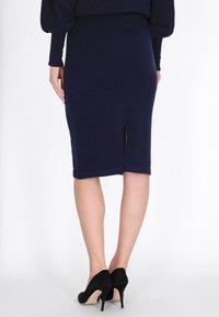 Fustă creion tricotată de culoare bleumarin, cu o fanta în spate, asortată cu pantofi cu toc înalt negri. Materialul pare moale, având o textură netedă.