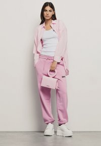 Camicia a righe rosa sopra una canottiera bianca, abbinata a pantaloni della tuta rosa e sneaker platform bianche. Tiene in mano una piccola borsa rosa.