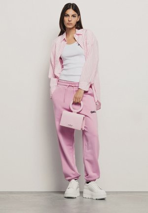 Camicia a righe rosa sopra una canottiera bianca, abbinata a pantaloni della tuta rosa e sneaker platform bianche. Tiene in mano una piccola borsa rosa.