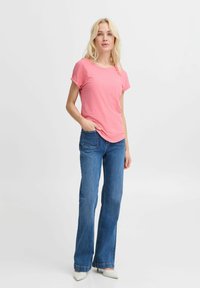 T-shirt rose à manches courtes avec un col rond, associée à un jean bleu évasé. Le modèle se tient devant un fond neutre, avec une tenue décontractée et des poches.