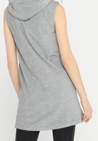 Bezrukawnik w odcieniu szarym, wykonany z miękkiego materiału, z kapturem, luźnym krojem i zaokrąglonym dołem. Idealny do warstwowego noszenia.