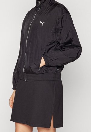 Person iført en sort Puma lynlåstrøje med elastiske manchetter og et sort skørt med en slids foran i siden.